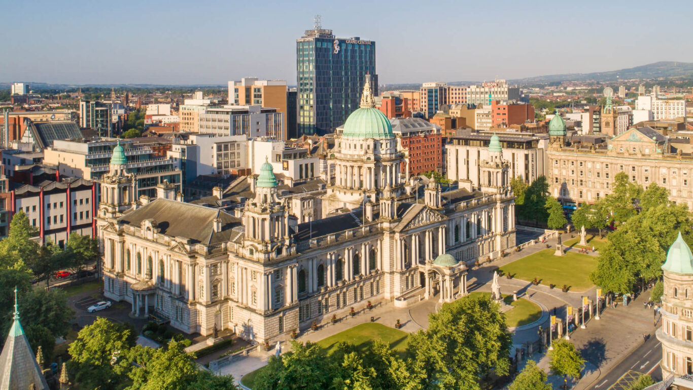 Aerial view of Belfast City Hall, a grand historic building with green domes, surrounded by lush trees in a bustling urban setting.