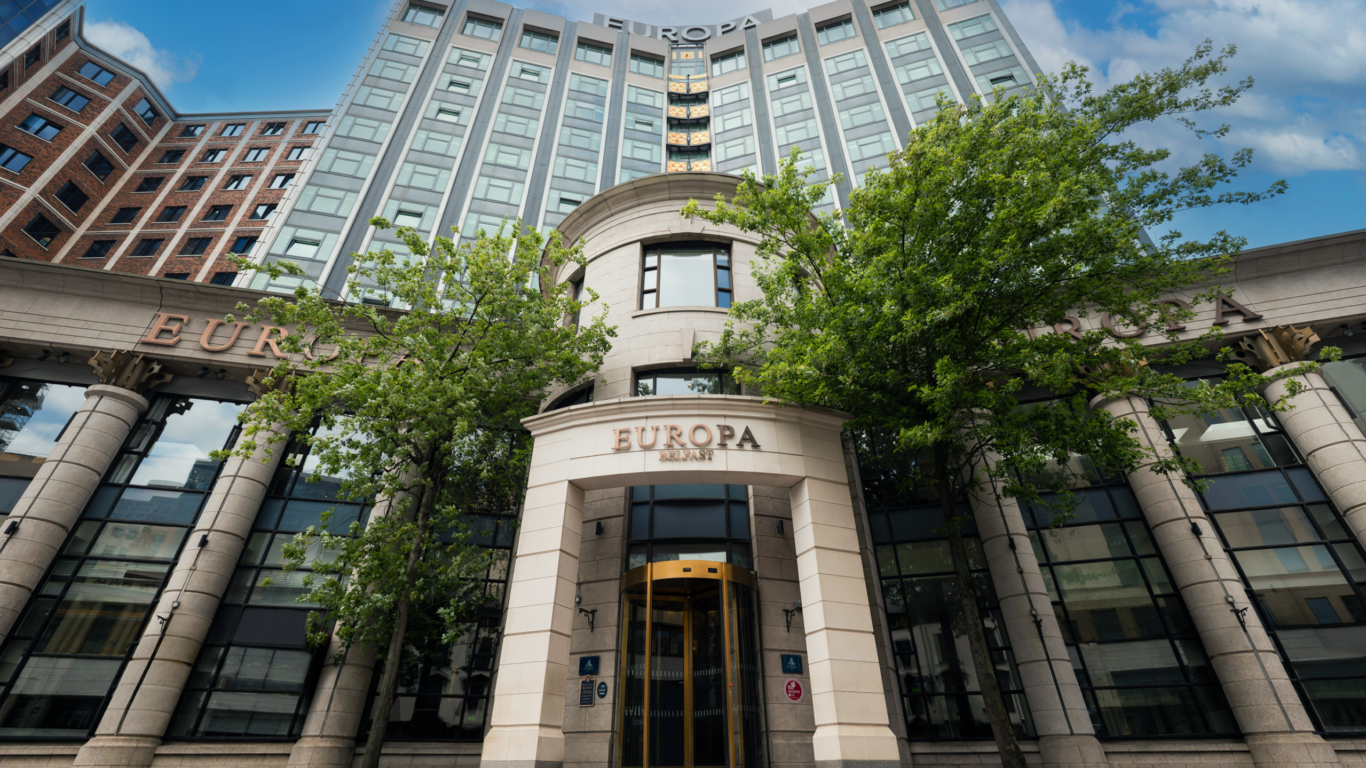 Grand hotel entrance with modern architecture, lush trees, and glass facade, inviting guests for a luxurious stay.