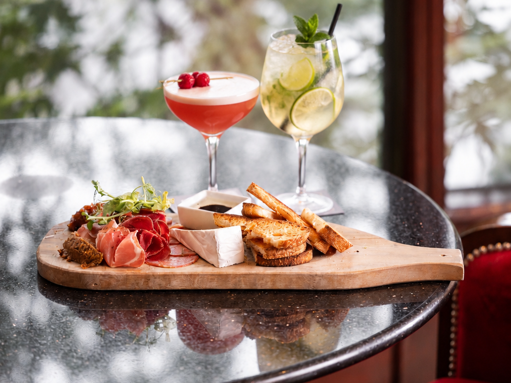 Elegant charcuterie board and cocktails on a glossy table, surrounded by plush seating, with a garden view.