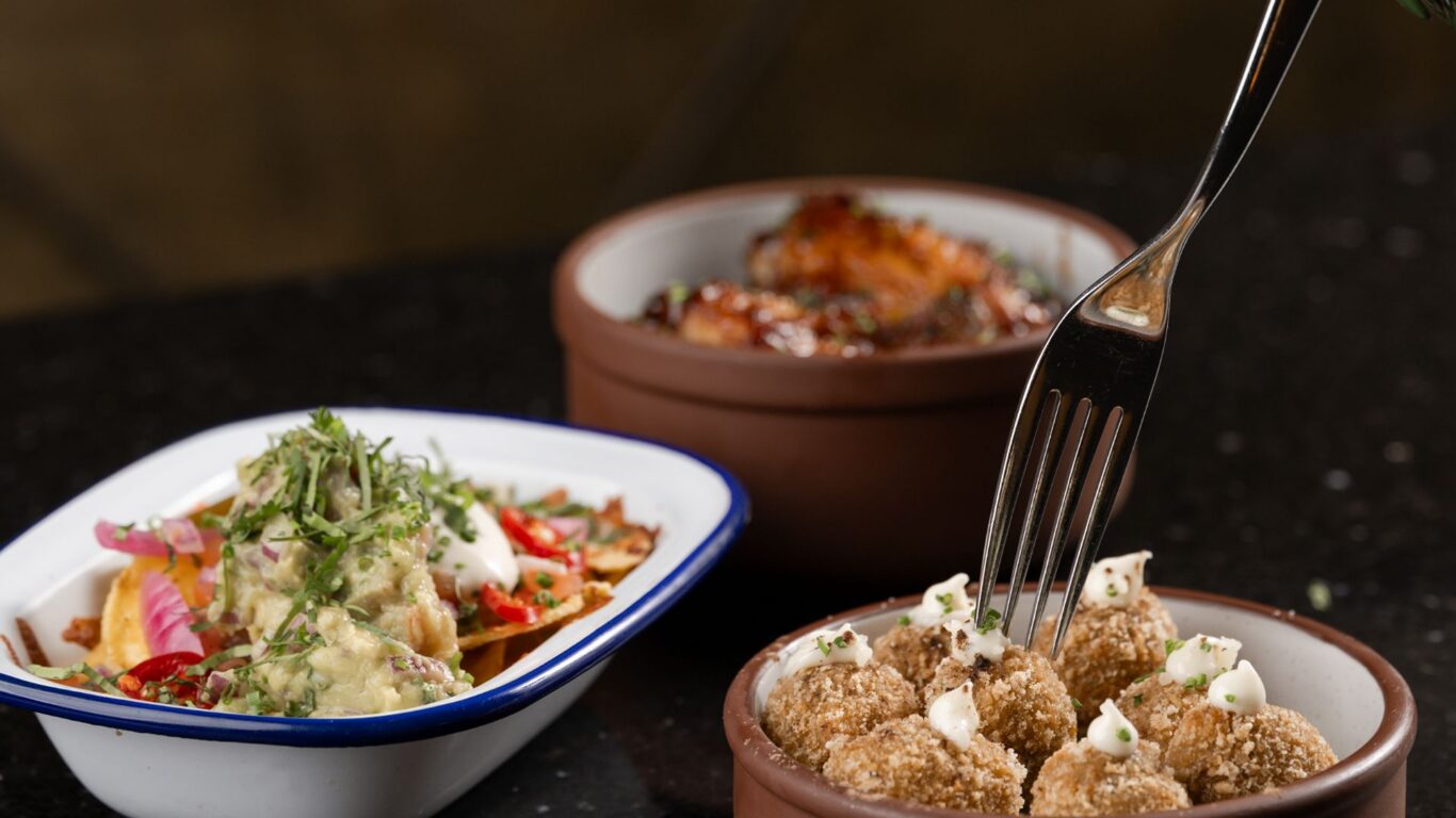 Three tapas dishes on a black granite bar &mdash; crispy croquettes with aioli, nachos with guacamole and pickled onions, and baked chorizo in a ramekin, fork poised to taste