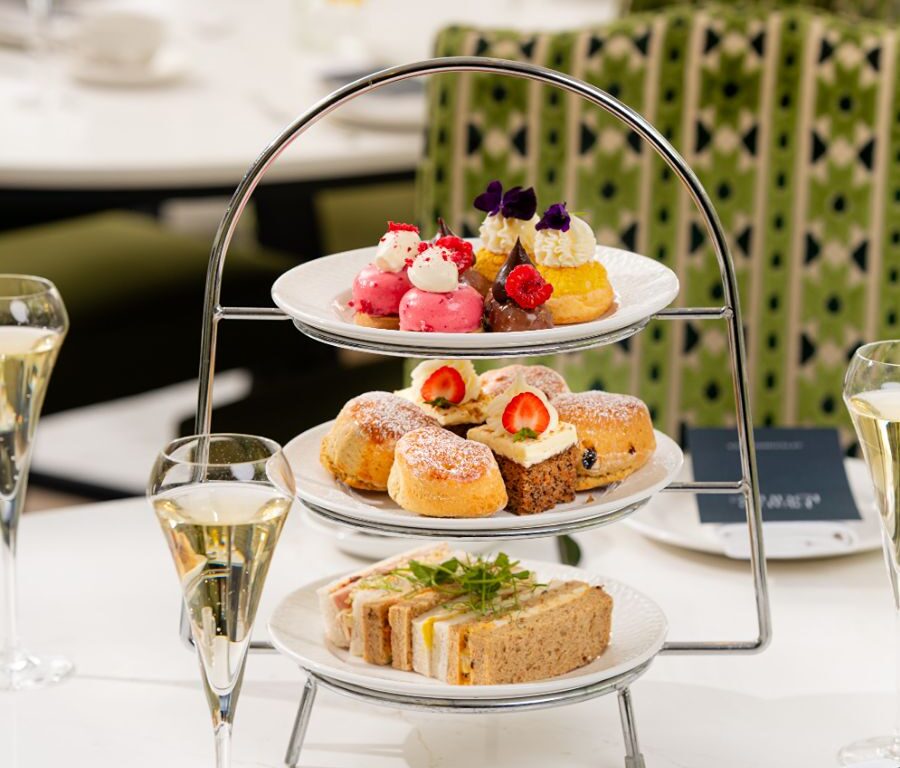 Three-tier afternoon tea stand with finger sandwiches, scones with cream and jam, assorted petit desserts, and champagne flutes in a bright hotel lounge