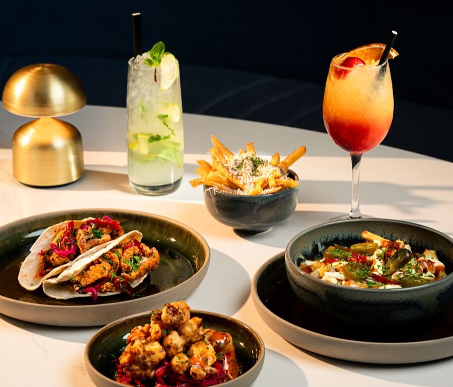 Gourmet sharing plates on a marble table — tacos, crispy cauliflower, loaded fries and pasta with two cocktails, modern hotel bar dining