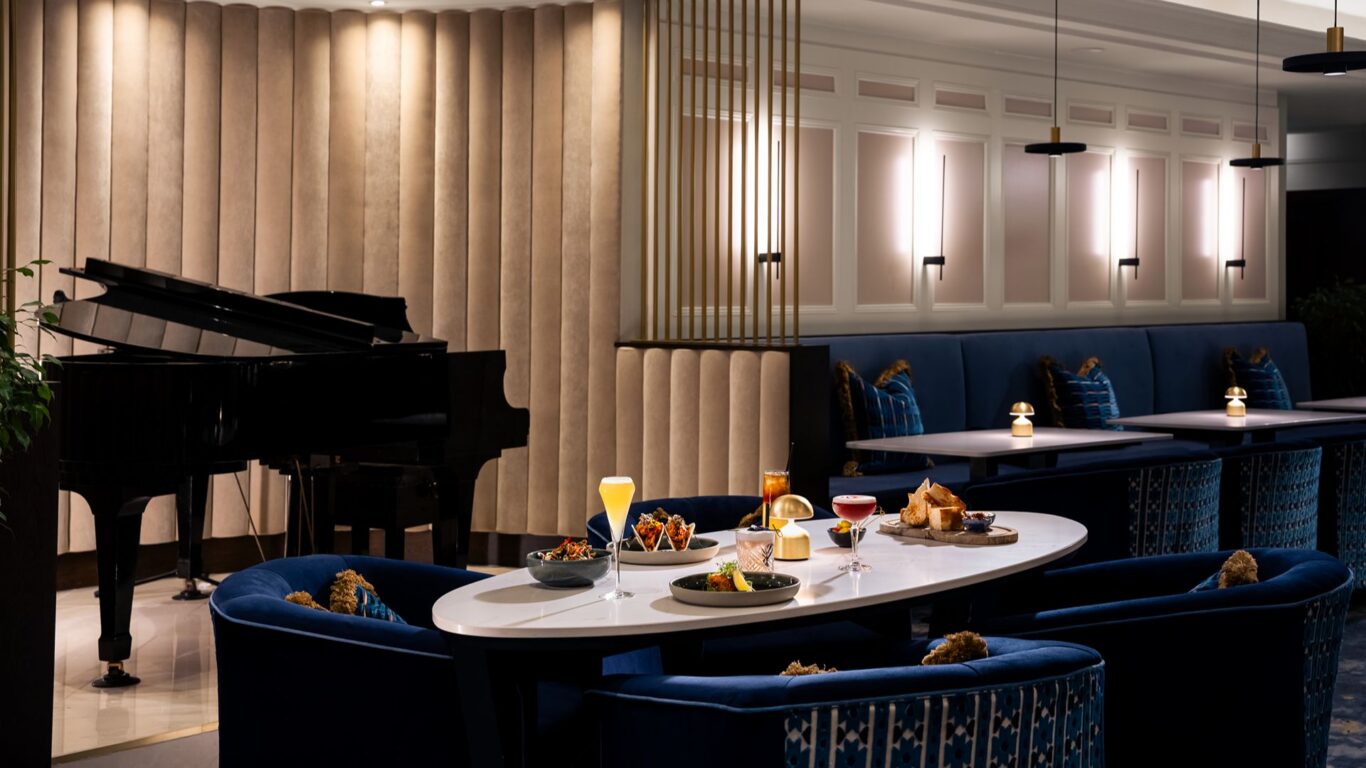 Chic hotel lounge with navy velvet seating, white oval table set with cocktails and tapas, grand piano and mood lighting